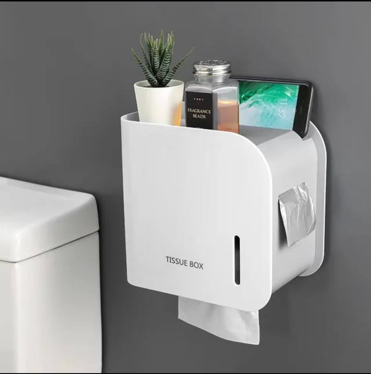Wall-mounted toilet paper holder with a white finish, featuring a slot for toilet paper and a shelf for other bathroom supplies, such as a mobile phone and a plant.